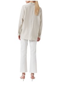 Chemise beige clair à manches longues avec une couture arrière, associée à un pantalon blanc ajusté et des talons blancs à brides, mettant en valeur une silhouette élégante.