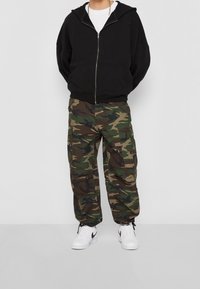 Felpa nera con zip, pantaloni cargo mimetici con più tasche e sneakers bianche con dettagli neri. Vestibilità rilassata con un look informale.