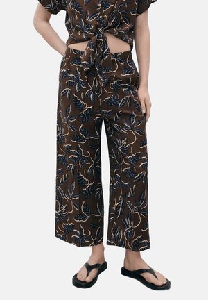 Pantalon marron à jambes larges avec imprimé de feuilles tropicales bleues et blanches, porté avec un crop top noué assorti et des sandales à plateforme noires.