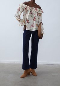 Blouse fleurie à épaules dénudées avec volants, associée à un jean évasé bleu foncé et des bottines à talons carrés marron, tenue avec une pochette beige.