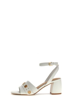 Sandalia blanca con tacón bloque, tiras en el tobillo y tachuelas decorativas doradas en la amplia tira frontal, punta abierta y cierre de hebilla.