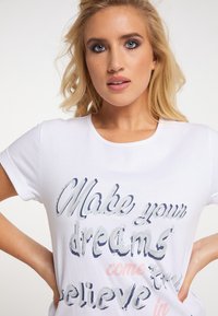 Weißes T-Shirt mit kurzen Ärmeln, das einen Text in verschiedenen Blau- und Rosatönen zeigt: "Mach deine Träume wahr, glaub daran." Weiche, lockere Passform.