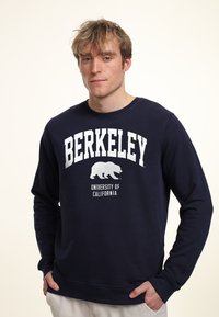 Felpa navy con la scritta "BERKELEY" e "UNIVERSITY OF CALIFORNIA" in bianco, accompagnata da una grafica di un orso sotto. Tessuto morbido e scollatura a girocollo.