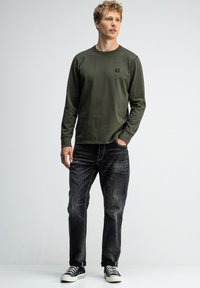Junger Mann steht mit der Hand in der Tasche, trägt ein olivgrünes Langarmhemd, verbaute schwarze Jeans und schwarze Sneakers vor einem weißen Hintergrund.