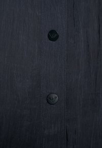 Dark gray garment featuring a textured fabric with two black buttons, one displaying a logo. Vertical seam details are visible.