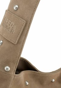 Zachte beige suede schoudertas met een textuurafwerking. Voorzien van metalen studs en een reliëf logo op de schouderband. Gebogen bovenopening.
