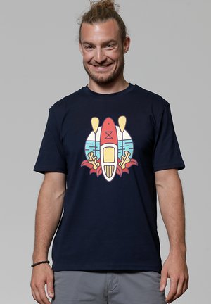 Mann mit zurückgebundenem Haar trägt ein marineblaues T-Shirt mit einem bunten Motiv eines Kajaks, Paddeln und einem wasserbezogenen Design.