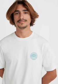 Camiseta de algodón blanca con un logo circular azul con diseño de sol y ola en el lado izquierdo del pecho. Mangas cortas y cuello redondo.