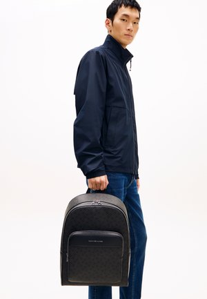 Mochila negra con un patrón texturizado, cierre de cremallera y bolsillo frontal. Llevada por una persona con chaqueta azul marino y vaqueros azules.