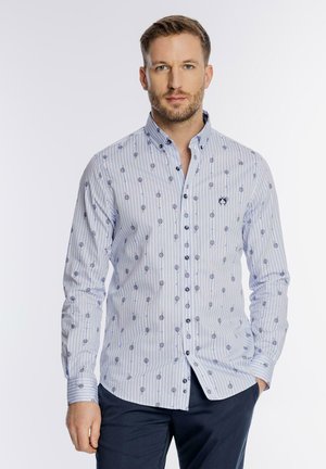 Mężczyzna ubrany w jasnoniebieską koszulę w paski z ciemnymi okrągłymi wzorami i granatowe spodnie, stojący z jedną ręką w kieszeni.