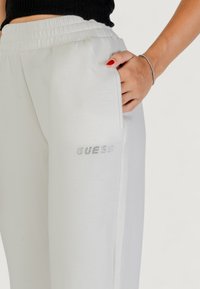 Un pantalon de survêtement gris clair avec une taille côtelée, des poches latérales et un logo "GUESS" ornée de cristaux sur la jambe gauche. Texture douce et lisse.