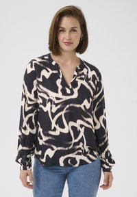 Mønstret bluse i sort og creme med lange ærmer, V-udskæring og afslappet pasform, stoppet ned i blå denimjeans. Blød stoftekstur.