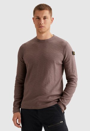 Hombre vistiendo un suéter marrón de manga larga y pantalones negros con bolsillos con cierre, de pie contra un fondo claro y liso.