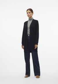 Cardigan lungo nero con design aperto, texture morbida, abbinato a un dolcevita grigio e jeans neri a gamba larga, indossato con scarpe slip-on color tan.