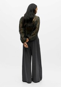 Dark green sheer lace blouse with long sleeves and floral patterns, paired with loose grey trousers. The model stands with hands behind back.