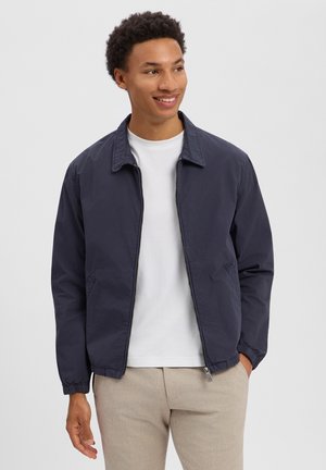 Uśmiechający się młody mężczyzna w granatowej kurtce na zamek błyskawiczny, białej koszulce i beżowych spodniach, stojący z rozluźnionymi rękami, z jedną w kieszeni.