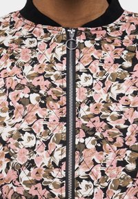 Κοντινό πλάνο μπουφάν με λουλουδάτο μοτίβο με ροζ, λευκά και καφέ λουλούδια και κεντρικό ασημί φερμουάρ με στρογγυλό δαχτυλίδι έλξης.