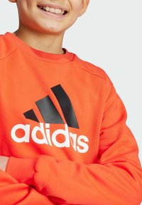 Sudadera naranja con el logo negro de Adidas y texto blanco. Presenta un cuello y puños acanalados, tejido de peso medio y corte relajado.
