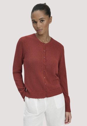 Vrouw met donker haar in een knot, gekleed in een roestkleurige, met knopen gesloten cardigan en witte broek, staand tegen een lichtgrijze achtergrond.