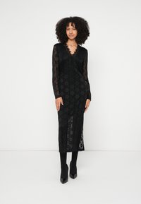 Crna čipkana haljina s dugim rukavima, V-izrezom i složenim uzorcima; duljine do koljena s bočnim prorezom; stilizirana s crnim visokim potpeticama.