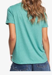 T-shirt teal a maniche corte con una vestibilità rilassata e una texture morbida, abbinato a pantaloni corti in denim. Etichetta visibile vicino all'orlo.