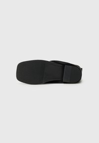 Semelle d'une chaussure noire à bout carré et talon bas présentée sur un fond gris clair.