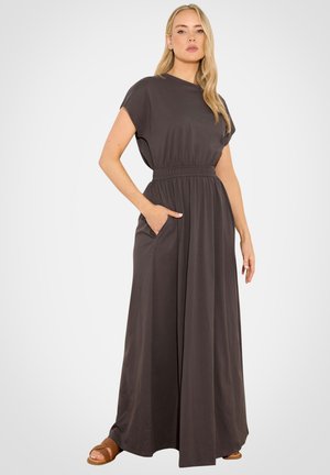 Vrouw staand met één hand in de zak, gekleed in een donkergrijze maxi-jurk met korte mouwen en bruine geweven sandalen, tegen een effen achtergrond.