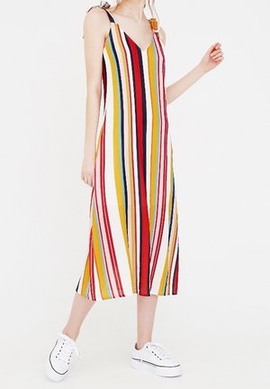 Kobieta ubrana w midi sukienkę bez rękawów z dekoltem w serek, w pionowe paski w kolorach czerwonym, żółtym, białym, czarnym i niebieskim, do tego białe trampki na platformie.