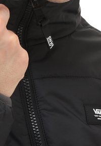 Giacca nera isolata con colletto alto, zip visibile con tirante e patch con logo sul petto. Superficie liscia e lucida.