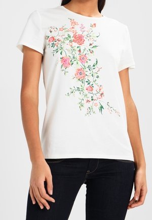 Biała koszulka z krótkim rękawem z różowym i koralowym kwiatowym wzorem układającym się ukośnie na przodzie, noszona z ciemnoniebieskimi dżinsami.