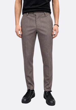 Uomo che indossa pantaloni eleganti marroni slim-fit, t-shirt nera a girocollo, braccialetto nero e scarpe stringate nere in piedi davanti a uno sfondo semplice.