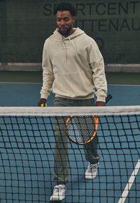 Light beige hoodie with front pocket, olive green sweatpants, and white athletic shoes; holding a yellow tennis ball and racket.