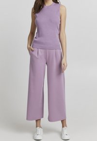 Liliowa bezrękawnikowa bluzka z okrągłym dekoltem w połączeniu z szerokimi liliowymi spodniami. Modelka ma na sobie białe sneakersy, ręce w kieszeniach.