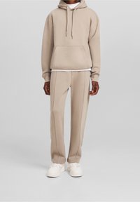 Felpa beige con tasca frontale e coulisse regolabili, abbinata a pantaloni affusolati coordinati, tessuto testurizzato e sneakers bianche.