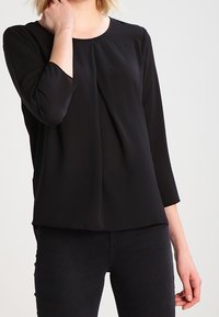 Blusa nera in tessuto leggero con scollo rotondo, maniche a tre quarti e dettagli con plissettatura frontale. Indossata con pantaloni neri.
