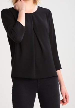 Blouse noire en tissu léger avec un col rond, des manches trois-quarts et des détails de plis sur le devant. Portée avec un pantalon noir.
