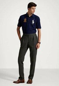 Νεαρός άνδρας φοράει ναυτικό πόλο με κεντημένα λογότυπα, γκρι πλισέ παντελόνια, καφέ δερμάτινα παπούτσια και ρολόι χειρός, στέκεται με τα χέρια στις τσέπες.