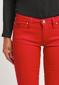 Czerwone dopasowane dżinsy o gładkiej fakturze, pięć kieszeni, z zamkiem błyskawicznym. Podkreślone małym, paskowanym detalem na przedniej kieszeni.