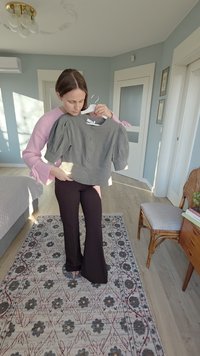 Blouse grise à manches courtes bouffantes, avec des boutons proéminents sur le devant. Portée en tenant le vêtement.