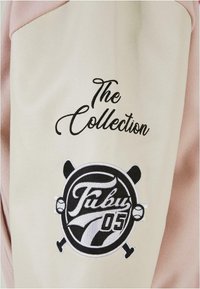 Krem- og rosa stoff med svart brodert tekst: "The Collection" og logo med "Fubu 05" og kryssede greiner. Glatt tekstur.