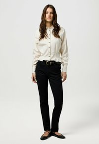 Camicia bianca con bottoni neri, abbinata a pantaloni neri a gamba dritta e scarpe basse, su uno sfondo neutro.