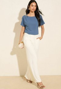 Top blu corto con maniche a sbuffo e dettaglio decorativo sulle spalle, abbinato a pantaloni bianchi a gamba larga e sandali con tacco nudi. Texture lisce in tutto l'outfit.