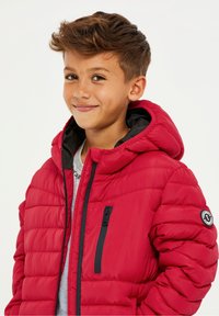 Veste rembourrée rouge avec capuche, dotée d'accents noirs, d'une poche poitrine zippée et d'un patch logo sur la manche. Texture lisse.