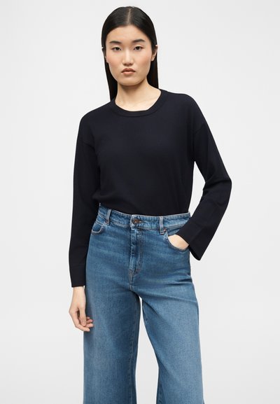 Νεαρή γυναίκα με ίσια σκουρόχρωμα μαλλιά, φοράει μαύρη μπλούζα με μακριά μανίκια και ψηλόμεσο μπλε τζιν, στέκεται με το ένα χέρι στην τσέπη.