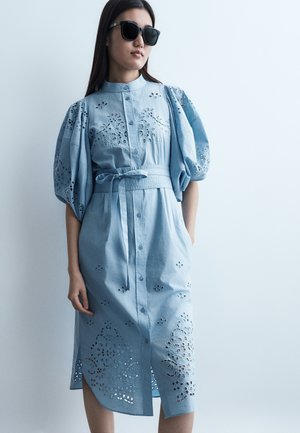 Γυναίκα που φοράει ένα ανοιχτό μπλε midi φόρεμα με κουμπιά, ζώνη, φουσκωτά μανίκια και κεντήματα με λουλουδάτο μοτίβο, συνδυασμένο με μαύρα γυαλιά ηλίου.