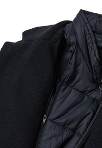 Giacca nera con uno strato esterno strutturato e una fodera interna liscia e isolante. Presenta una chiusura con cerniera e un colletto rialzato.