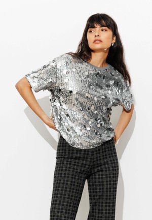 T-shirt à paillettes argentées avec manches courtes, col rond et coupe ample. Associé à un pantalon à carreaux noir. Fond uni.