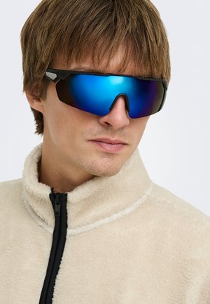 Jeune homme portant des lunettes de soleil de sport bleues réfléchissantes et une veste polaire couleur crème avec une fermeture éclair noire.