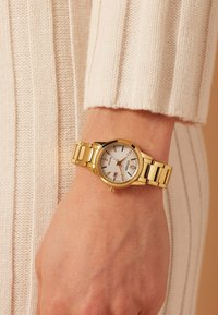 Gouden metalen polshorloge met een ronde witte wijzerplaat, uurmarkeringen en datumaanduiding. Bevestigd aan een gepolijste metalen armband, getoond aan een hand.