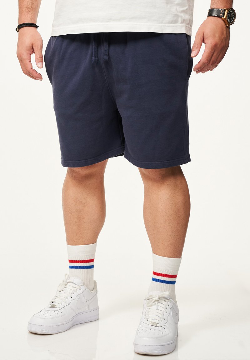 Dunkelblaue Baumwollshorts mit elastischem Bund und Kordelzug. Kombiniert mit weißen Turnschuhen und weißen Socken mit roten und blauen Streifen.